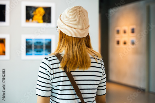 Asian woman standing she looking art gallery in front of colorful framed paintings pictures on white wall, young female watch at photo frame to leaning against at show exhibition gallery, Back view photo