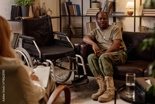Black veteran with disability sitting on couch in relaxed pose and talking to therapist taking notes © AnnaStills