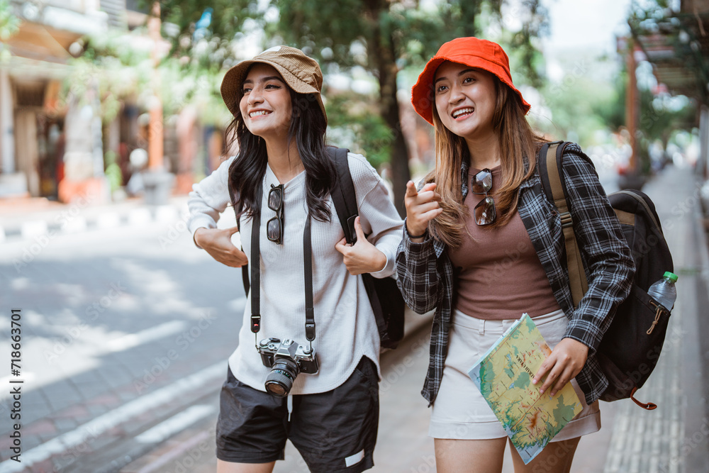 Fototapeta premium two backpacker girls walk to find a tourist destination carrying a map while on the roadside