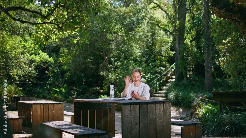 Fototapeta premium Beautiful woman greeting smartphone camera sitting wooden table in green park. 