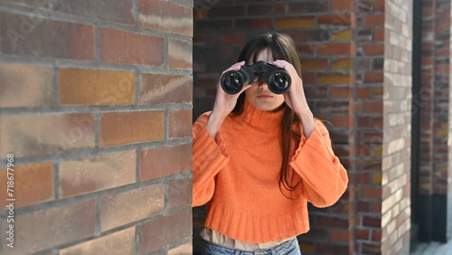 A young woman peeks around the corner of a building and spies on someone with binoculars
