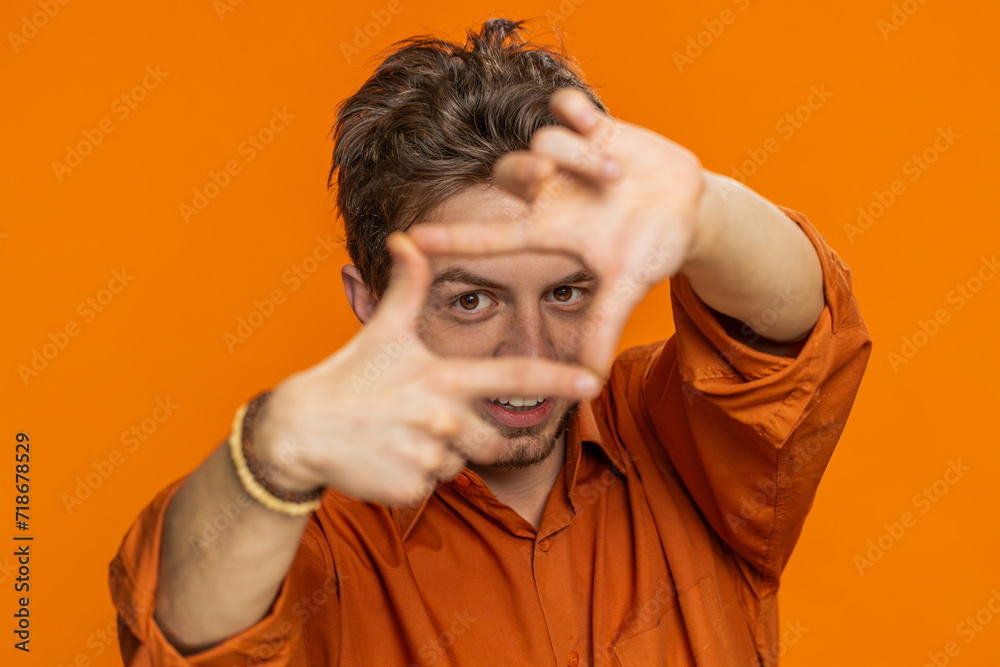Caucasian man photographer gesturing picture frame with hands, looks through fingers and focusing on interesting moment, imitating zoom and cropping nice image. Young guy isolated on orange background Caucasian man photographer gesturing picture frame with hands, looks through fingers and focusing on interesting moment, imitating zoom and cropping nice image. Young guy isolated on orange background