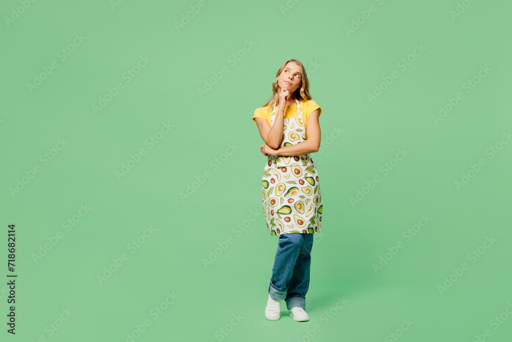 Full body smiling happy fun young housewife housekeeper chef cook baker woman wear apron yellow t-shirt prop up chin look aside on area isolated on plain green background studio. Cooking food concept. Full body smiling happy fun young housewife housekeeper chef cook baker woman wear apron yellow t-shirt prop up chin look aside on area isolated on plain green background studio. Cooking food concept.