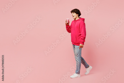 Full body side view young Caucasian man wears hoody casual clothes hold takeaway delivery craft paper brown cup coffee to go isolated on plain pastel light pink background studio. Lifestyle concept.
