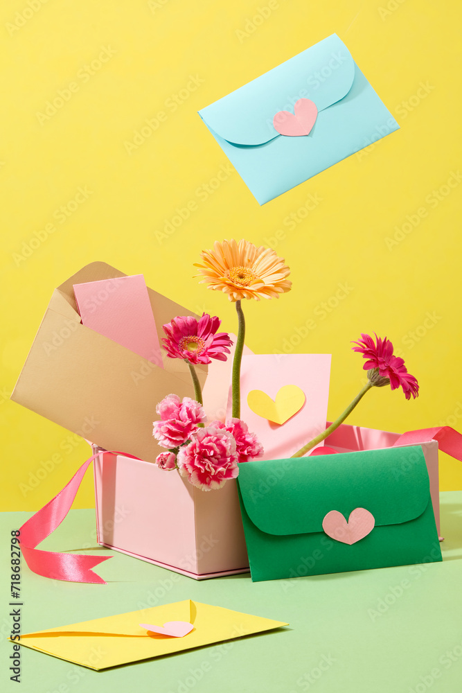 On a yellow background, fresh flowers with gerbera and carnation decorated with cute handmade envelopes on a pink gift box. Creative idea for women’s day decoration. Front view
