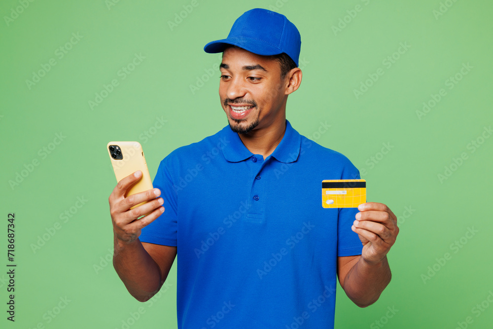 Professional delivery guy employee man wears blue cap t-shirt uniform workwear work as dealer courier hold credit bank card use mobile cell phone isolated on plain green background. Service concept. Professional delivery guy employee man wears blue cap t-shirt uniform workwear work as dealer courier hold credit bank card use mobile cell phone isolated on plain green background. Service concept.