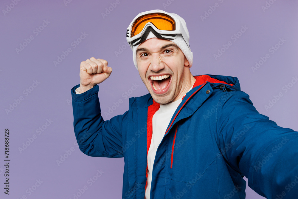 Close up skier man wear blue windbreaker jacket ski goggles mask hat do selfie shot on mobile cell phone do winner gesture spend weekend winter season in mountains isolated on plain purple background.