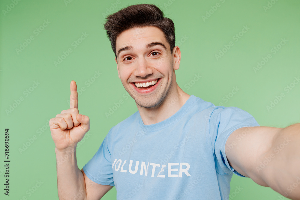 Young smart man wear blue t-shirt white title volunteer doing selfie shot pov mobile cell phone point finger up isolated on plain pastel green background. Voluntary free team work help grace concept. Young smart man wear blue t-shirt white title volunteer doing selfie shot pov mobile cell phone point finger up isolated on plain pastel green background. Voluntary free team work help grace concept.