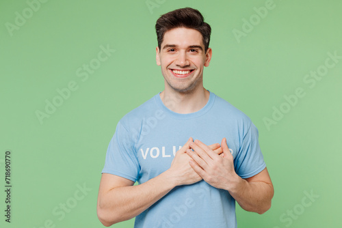 Young grateful happy man wears blue t-shirt white title volunteer put folded hands on heart isolated on plain pastel light green background. Voluntary free work assistance help charity grace concept. photo