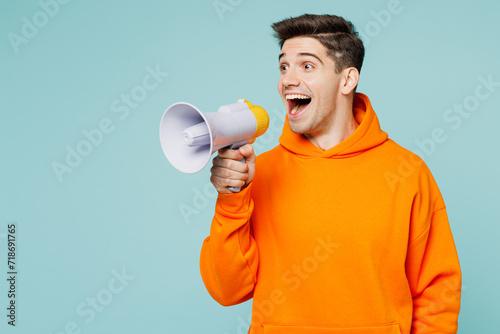 Young man wear orange hoody casual clothes hold in hand megaphone scream announces discounts sale Hurry up isolated on plain pastel light blue cyan color background studio portrait. Lifestyle concept