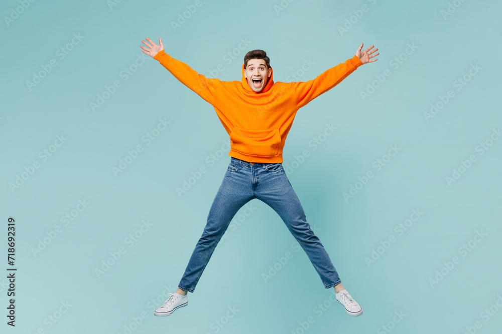 Full body young excited fun man wears orange hoody casual clothes jump high with outstretched arms hands isolated on plain pastel light blue cyan color background studio portrait. Lifestyle concept