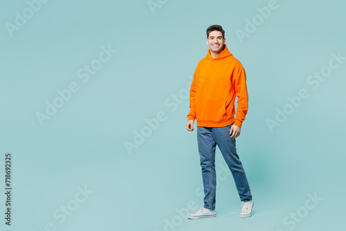 Full body smiling happy cheerful young man he wears orange hoody casual clothes look camera walking going isolated on plain pastel light blue cyan color background studio portrait. Lifestyle concept.