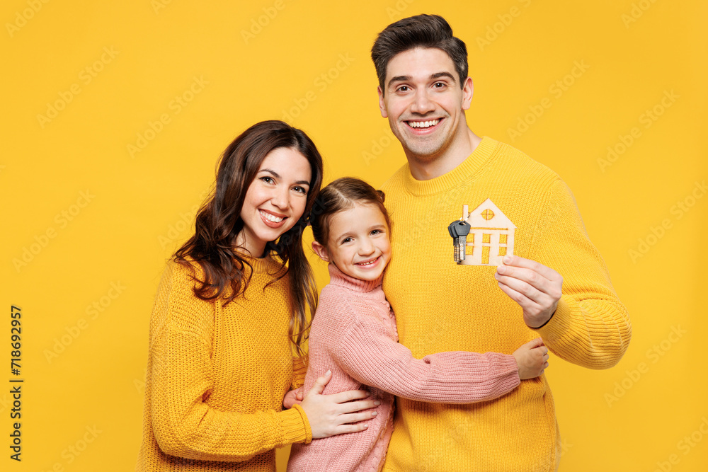 Fototapeta premium Young happy fun parents mom dad with child kid girl 7-8 years old wear pink sweater casual clothes hold keys and wooden house mockup hug cuddle isolated on plain yellow background. Family day concept.