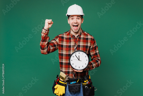 Young employee laborer man wear red shirt hardhat hat work hold clock do winner gesture isolated on plain green background. Instruments accessories for renovation apartment room. Repair home concept. photo