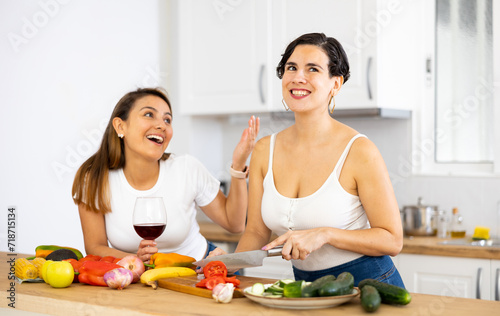 Smiling young Hispanic woman cooking with sister in home kitchen, chatting cheerfully and drinking wine while preparing vegetable salad. Happy family relationship concept..