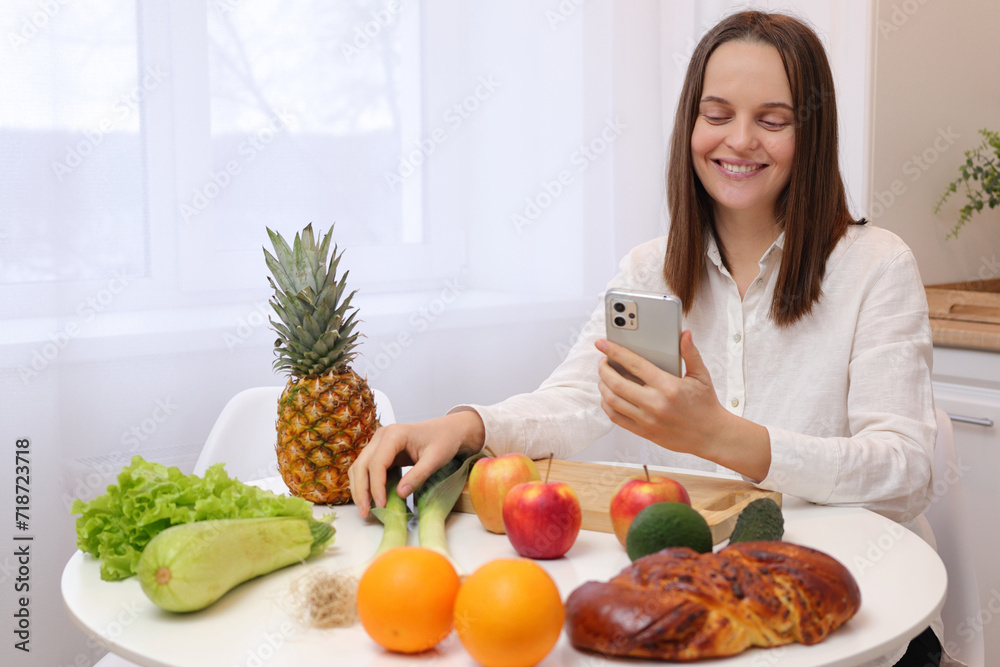 Smiling happy Caucasian brown haired woman sitting at kitchen table with fruit and vegetables preparing a delicious dinner with farm-fresh produce and scrolling online on mobile phone Smiling happy Caucasian brown haired woman sitting at kitchen table with fruit and vegetables preparing a delicious dinner with farm-fresh produce and scrolling online on mobile phone