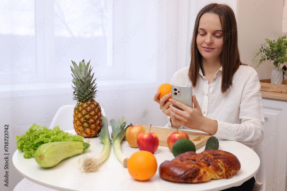 Beautiful attractive Caucasian brown haired woman sitting at kitchen table with fruit and vegetables using smartphone for browsing internet pages searching recipes of healthy organic meals