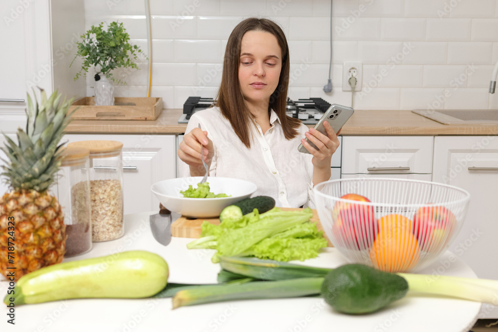 Confident Caucasian brown haired woman sitting at kitchen table with fruit and vegetables using smartphone while eating fresh green salad with lettuce leaves