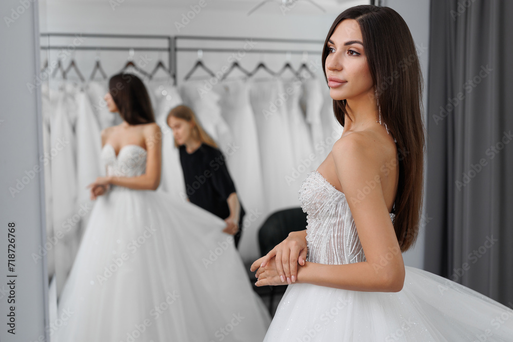 A portrait of a beautiful young bride who is trying on a white wedding dress in a store is reflected in the mirror.