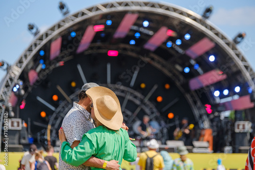A loving couple hugs and dancing enjoying the ambiance in front of a festival music stage on a sunny day.