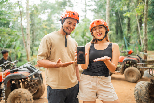 asian couple with savety equipment showing the blank phone to the camera while standing around the atv arena