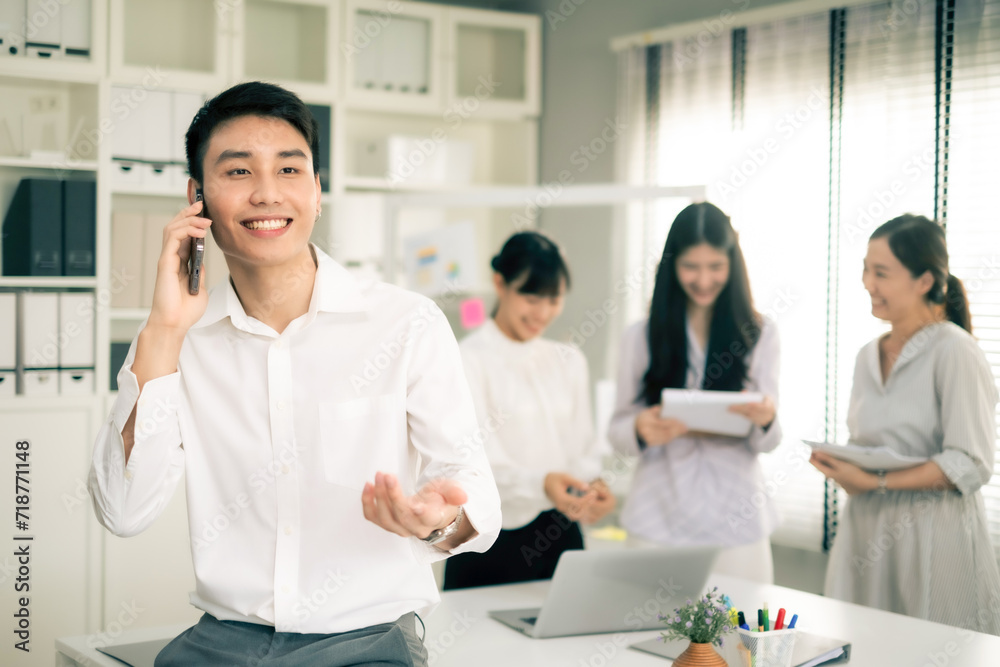 Young Asian business man using smartphone while sitting on table in office with smile. Man in white shirt looking away while talking to someone on mobilephone with co-worker background. Young Asian business man using smartphone while sitting on table in office with smile. Man in white shirt looking away while talking to someone on mobilephone with co-worker background.