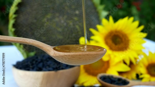 Liquid sunflower oil is poured into a spoon against a background of yellow flowers and sunflowers, and a wooden bowl filled with seeds stands nearby.