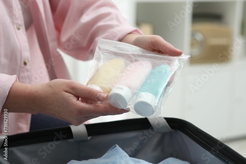 Woman holding plastic bag of cosmetic travel kit over suitcase indoors, closeup. Bath accessories