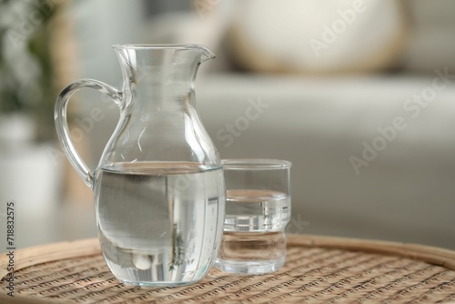 Jug and glass with clear water on wicker surface against blurred background, closeup