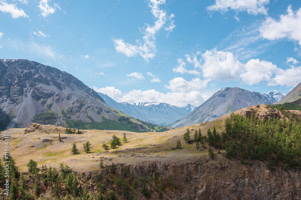 View above cliff to alpine green valley and large mountain range with snowy top far away. Sparse conifer trees on precipice edge in high mountains. Open coniferous forest on rocky wall above abyss. View above cliff to alpine green valley and large mountain range with snowy top far away. Sparse conifer trees on precipice edge in high mountains. Open coniferous forest on rocky wall above abyss.