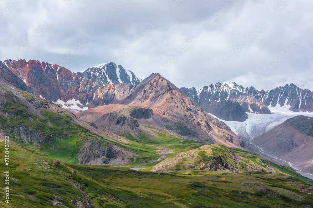 Vivid dramatic alpine scenery with green hills and rocks with view to rocky pointy peak, sharp rockies of red color, large snow mountain top, snowy range and big glacier tongue under gray cloudy sky.