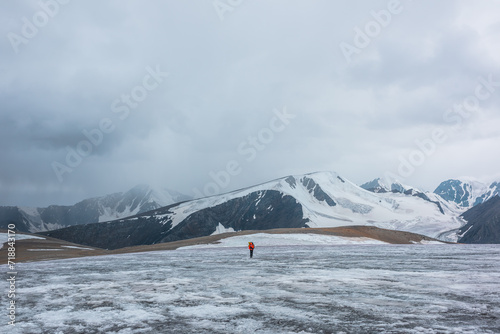 Man on ice pass with view to big glacier tongue among sharp rocks and large snow-capped mountains in rainy low clouds. Guy against dark atmospheric mountain silhouettes in rain in gray cloudy sky.