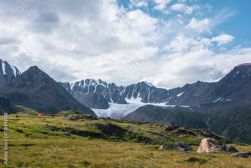 Dramatic scenery with orange tent in wide alpine valley among green hills and rocks with view to big glacier tongue and large snow-capped mountain range under clouds in blue sky in changeable weather. © Daniil