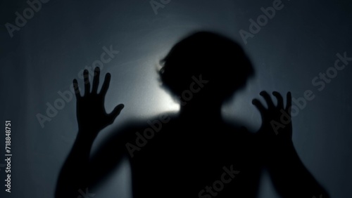 The dark silhouette of a man behind a matt curtain, illuminated by a beam of light. The man beats his fist, trying to break the invisible barrier. The concept of a ghost.