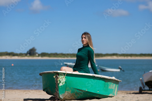 Young, beautiful blonde woman in an elegant green dress is sitting in a green fisherman's boat on the seashore. In the background the blue sea with the boats and the horizon. © @skuder_photographer