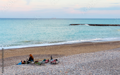 Travel at sea. People relax on stone beach by sea. View of coastline of pebble stone beach. Man and woman relax on beach after traveling bike ride. Cyclists resting after sports on sunset beach.