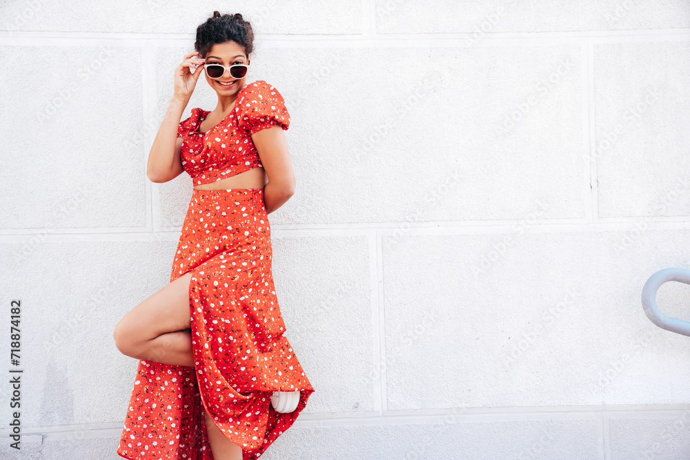 Young beautiful smiling hipster woman in trendy summer red dress. Carefree woman posing in the street near wall. Positive model outdoors in at sunny day. Cheerful and happy. In sunglasses Young beautiful smiling hipster woman in trendy summer red dress. Carefree woman posing in the street near wall. Positive model outdoors in at sunny day. Cheerful and happy. In sunglasses