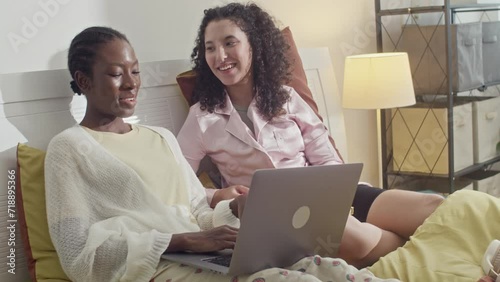 Medium shot of cheerful biracial girls chatting with friends on laptop while lying in bed at dormMedium shot of cheerful biracial girls chatting with friends on laptop while lying in bed at dorm
