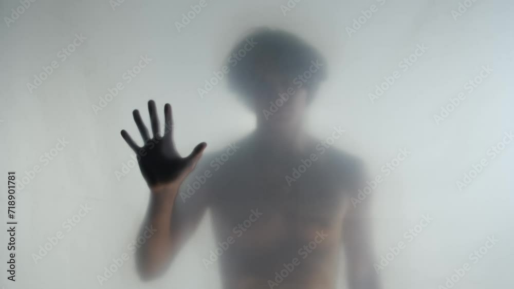 A male silhouette with a naked torso behind a transparent frosted curtain or glass close up. A man is running his hand over the surface of the curtain. A male silhouette with a naked torso behind a transparent frosted curtain or glass close up. A man is running his hand over the surface of the curtain.