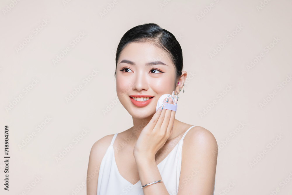 Young asian woman smile looking happy applying makeup with puff makeup on beige background.