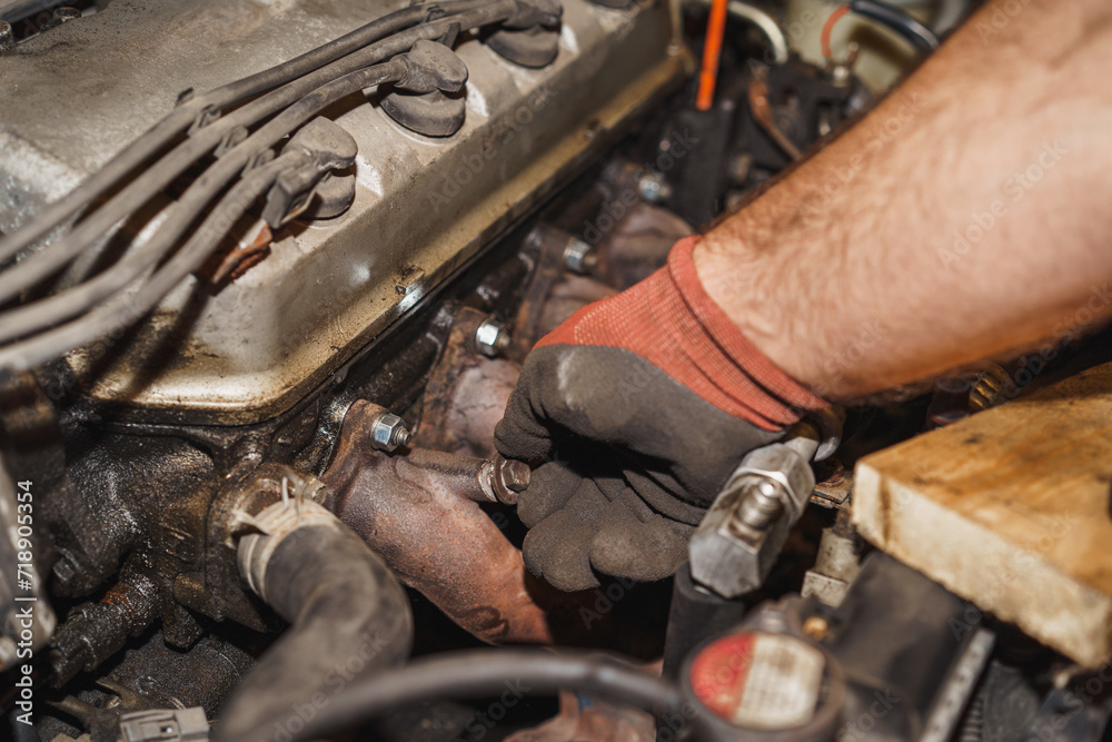 The mechanic is hand-tightening the bolt onto the exhaust pipe to reattach it to the car. Exhaust manifold, valve cover, spark plug cables, dipstick. The mechanic is hand-tightening the bolt onto the exhaust pipe to reattach it to the car. Exhaust manifold, valve cover, spark plug cables, dipstick.