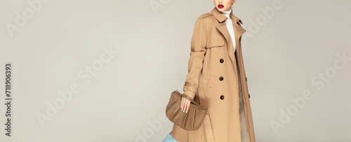 Autumn clothing shopping concept. Fashionable young model posing against grey background dressed in trench coat and holding stylish  bag. photo