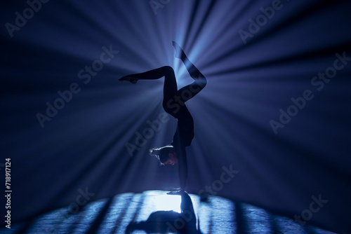 Female gymnast on dark background of studio with backlight. Acrobatic girl performing handstand. Modern choreography and acrobatics creative advertising concept. photo