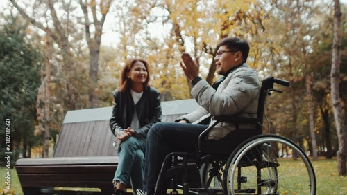 Disabled asian man in a wheelchair talking and gesturing with an asian woman on a bench in an autumn city park. Concept of acceptance, love and support for people with disability photo