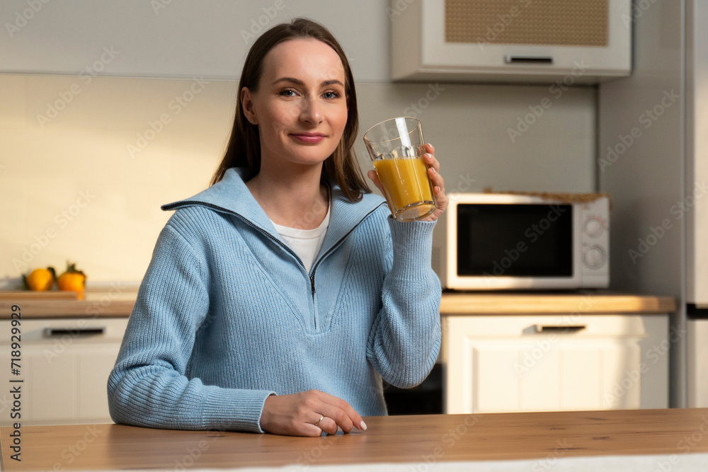 Young brunette dressed in a blue sweater drinks orange juice from a glass while sitting at the kitchen table. Young brunette dressed in a blue sweater drinks orange juice from a glass while sitting at the kitchen table.