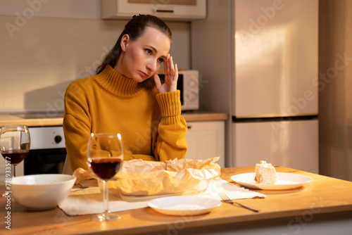 A contemplative woman sits at a table with a slice of cake and a glass of red wine in a warmly lit kitchen. A woman is crying alone. 