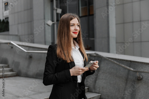 Young business woman wearing black dress code smiling answering message or chatting, using phone and looking at the camera