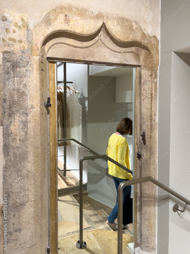 From the rear view, a woman gracefully descends a staircase in a luxury fashion store with a large mirror and a sculpted stone door entrance, creating an opulent interior.