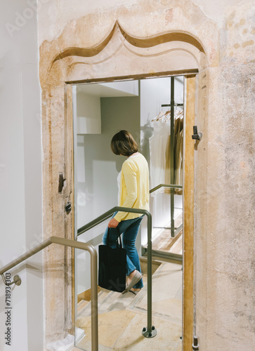 An unrecognizable woman stands on a staircase inside a modern retail fashion store, near an ancient door with a mirror, blending the old and new