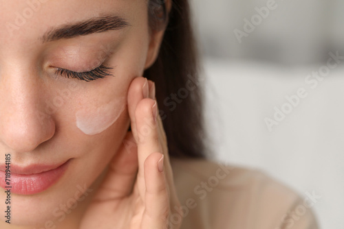 Young woman with dry skin applying cream onto her face against blurred background, closeup. Space for text